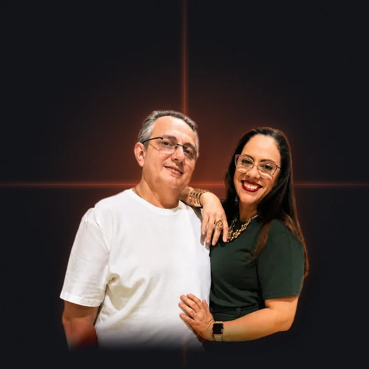 Pr. Júnior Bill e a Pastora Alessandra Mota abraçados e sorrindo, sobre fundo escuro com brilho terracota